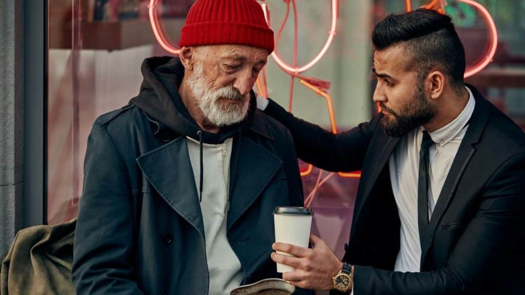 A man in a suit gives a cup to an older man in a red beanie and dark coat outside.