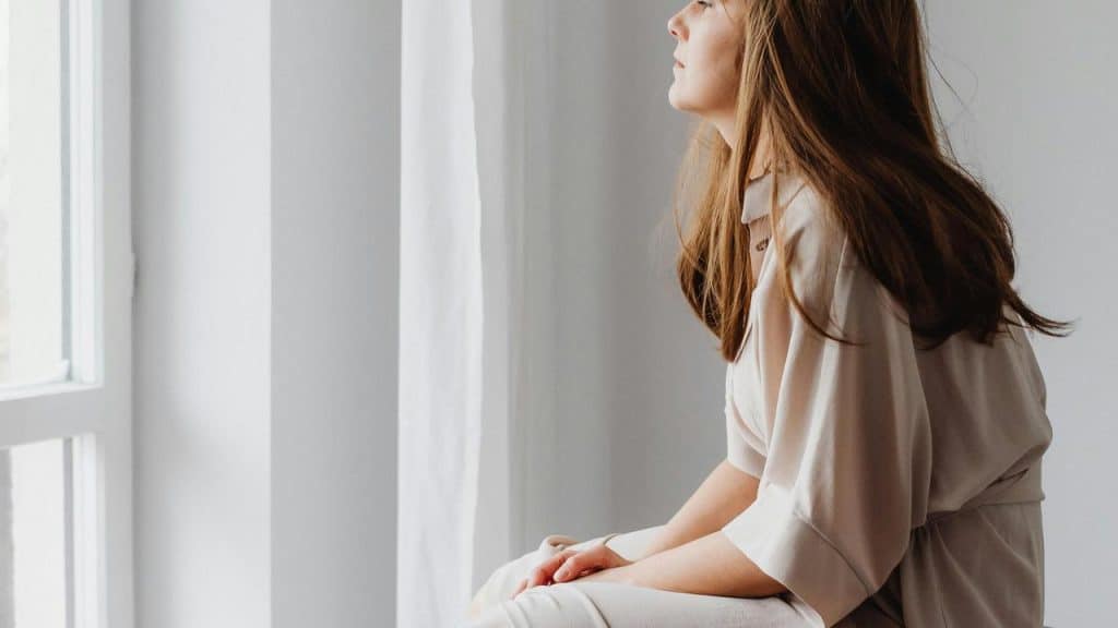 A woman in a light outfit sits by a window looking thoughtful.