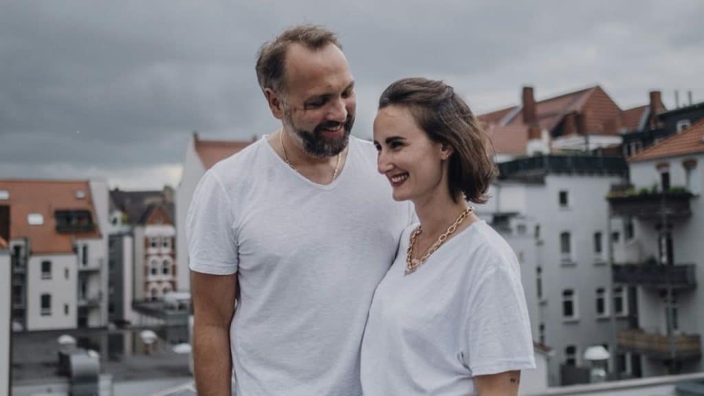 Smiling couple in matching white T-shirts and gold necklaces standing on a rooftop overlooking a town.