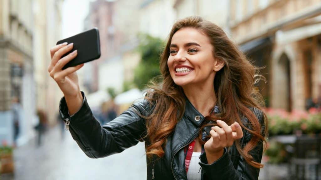 Smiling woman in leather jacket holding phone to take a selfie on a city street.