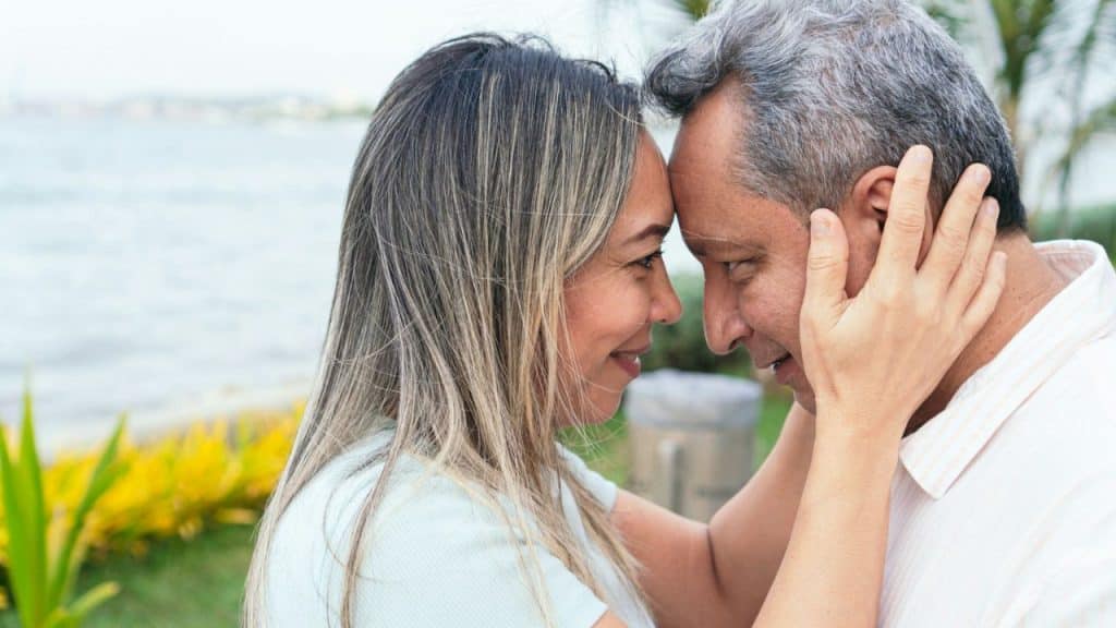 Middle-aged couple touching foreheads and smiling at each other outdoors by water.