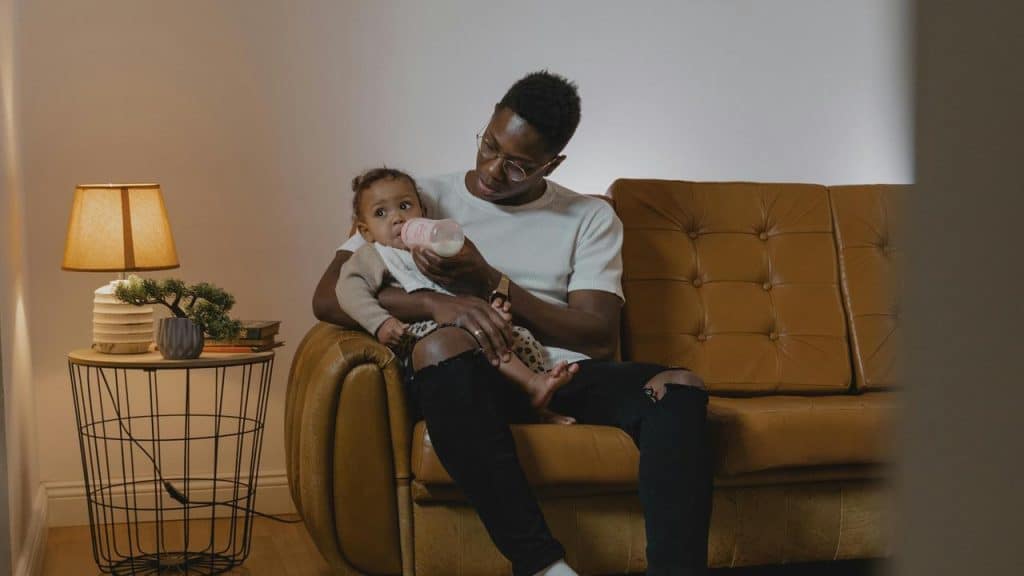A man sitting on a couch feeding a baby with a bottle.