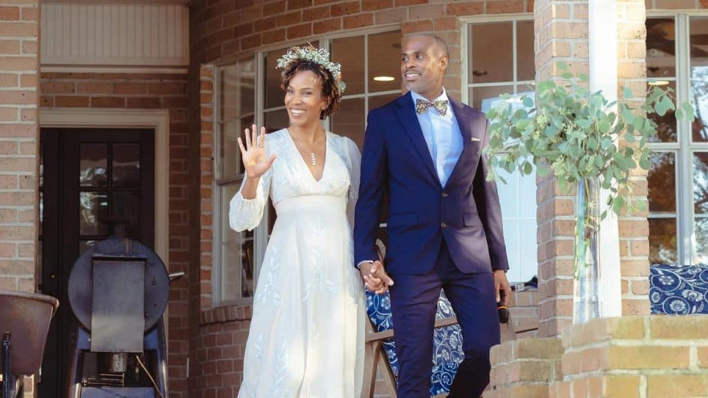 A smiling couple in wedding attire stands on a porch, holding hands and waving.