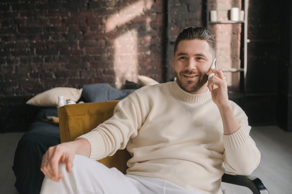 Bearded Man in White Knitted Sweater Sitting on Yellow Sofa Chair Using Cellphone