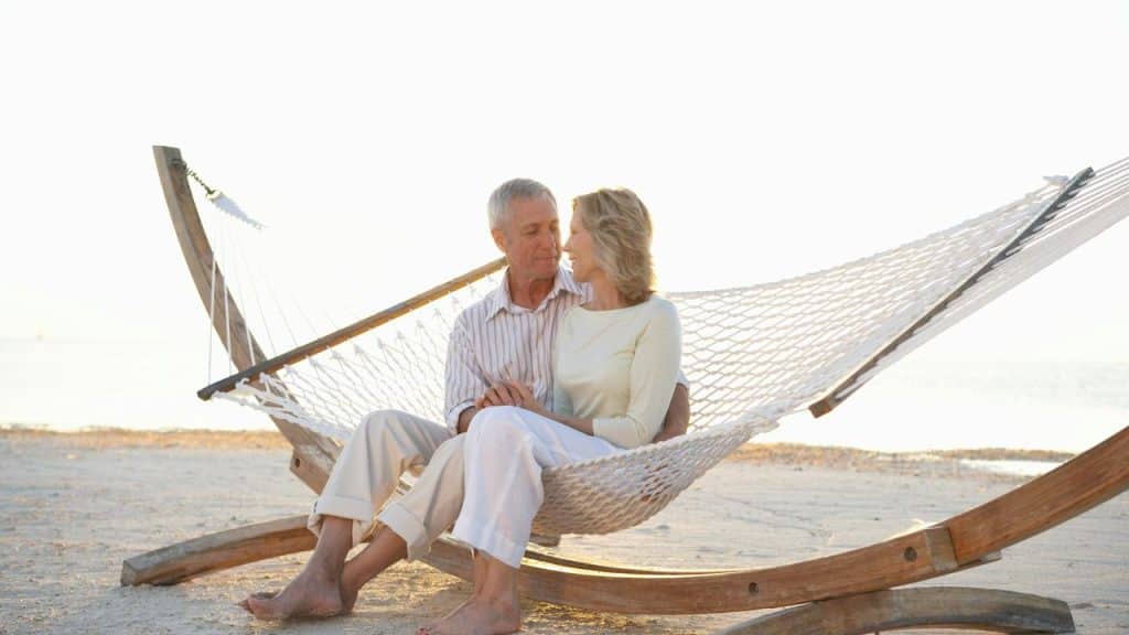 An older couple sits together on a hammock by the beach, looking at each other.