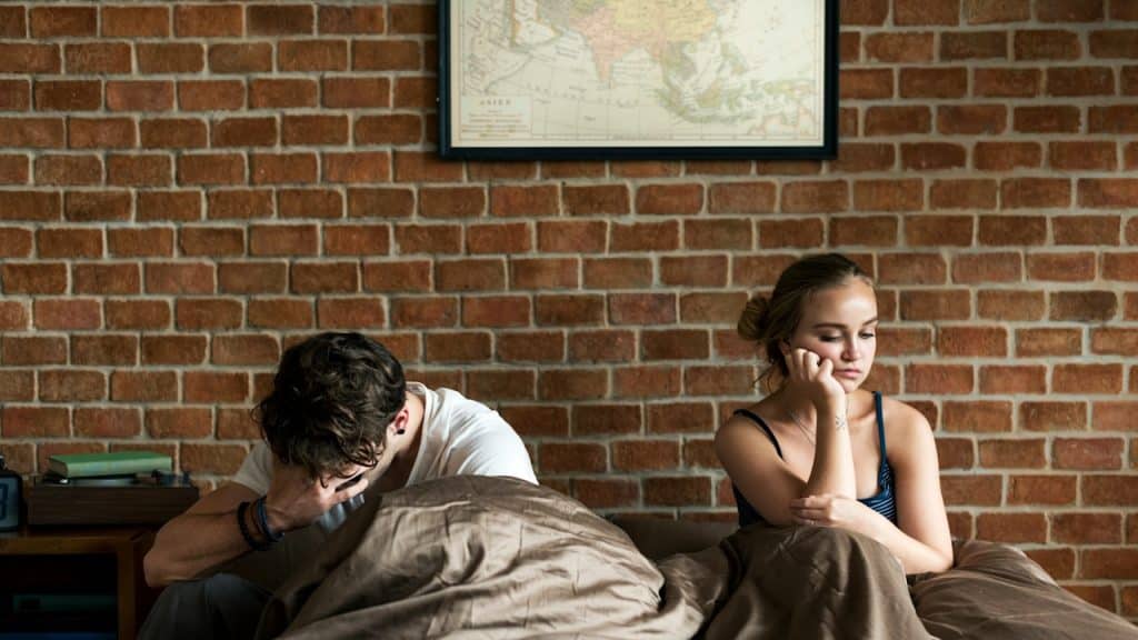 A man and a woman sit on opposite sides of a bed with their backs to a brick wall, both looking unhappy.