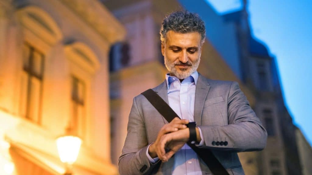 A bearded man in a suit looking at his smartwatch on a city street at night.
