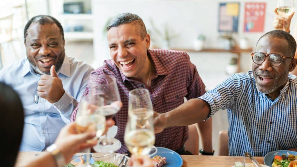Three men smiling and laughing while toasting with glasses at a table.