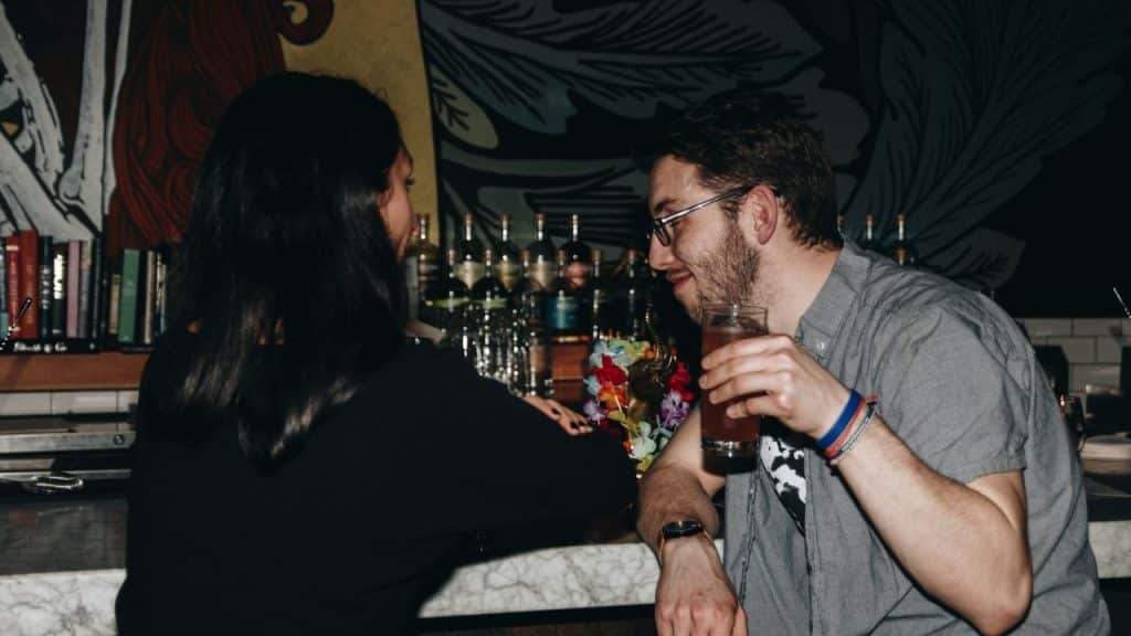 A smiling man holding a drink talks to a woman across a bar counter in a dimly lit setting.