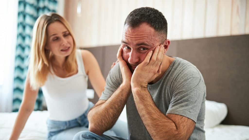 A distraught man sits on a bed, holding his head in his hands, while a concerned woman talks to him.