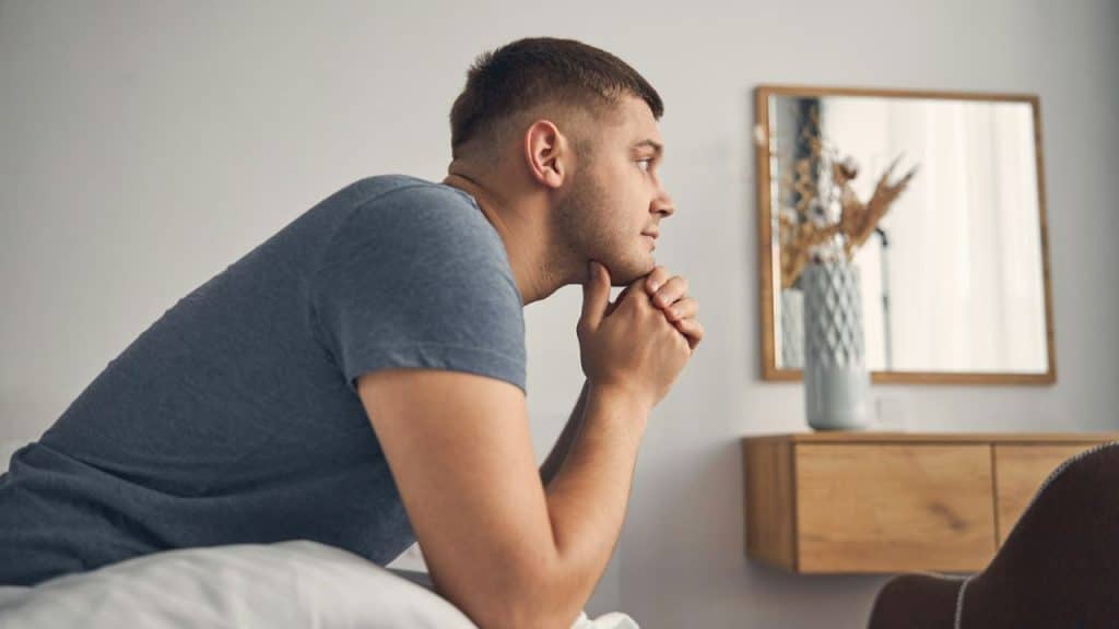 A contemplative man sits on a bed, looking off-camera with his hands clasped.