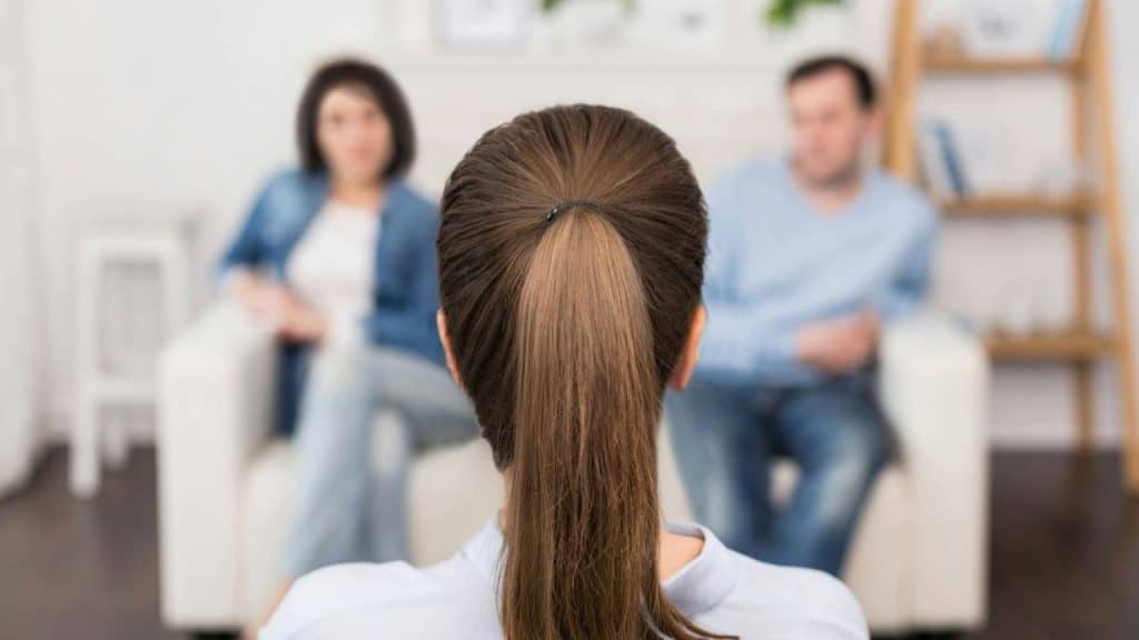 A therapist with her back to the camera sits across from a man and woman on a couch.