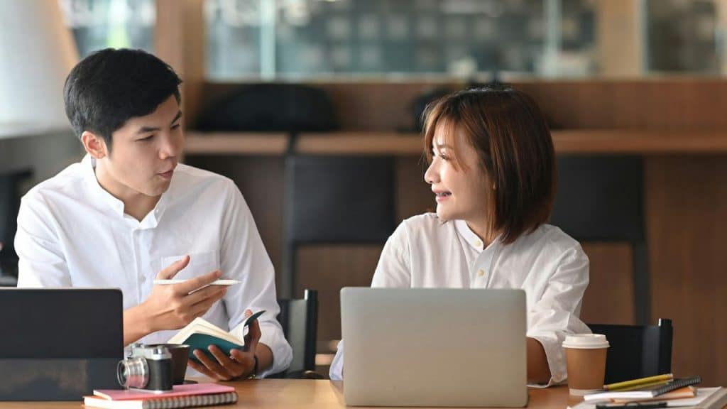 Two young professionals in white button-down shirts are sitting at a table with a laptop, talking and holding a notebook.