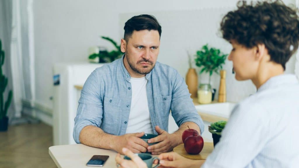 A stern-faced man in a blue button-down shirt sits across a table from a woman with curly hair, holding a coffee cup.