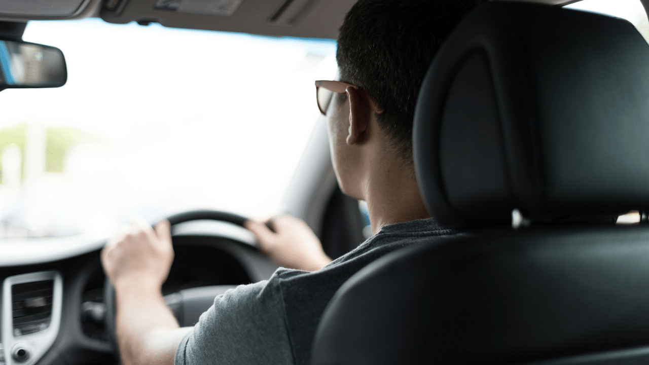 View from the back seat of a driver wearing sunglasses and holding the steering wheel.