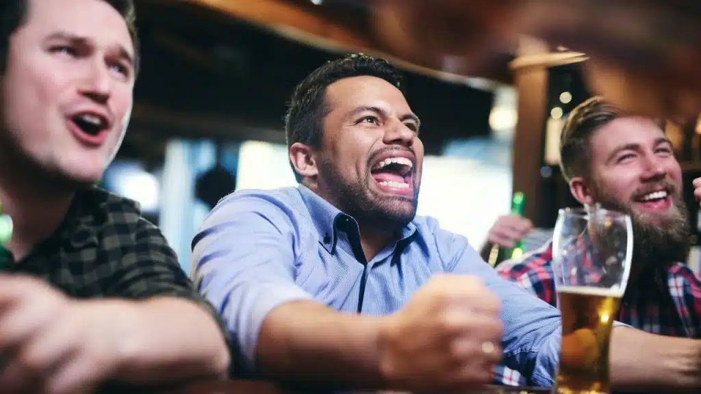 Three expressive men are enthusiastically watching a game with a beer at a bar.