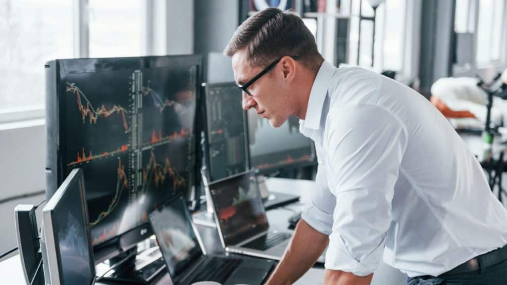 Man in a white shirt and glasses looking intently at multiple computer screens displaying stock charts.