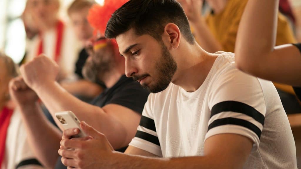 A focused young man with a beard and a white t-shirt with black stripes on the sleeves looks down at his smartphone while sitting in a crowd of cheering people at an event or game.
