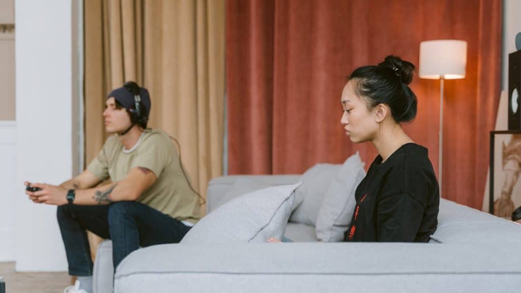 A young man is absorbed in playing a video game on the left, while a young woman sits looking away with a troubled expression on the right.