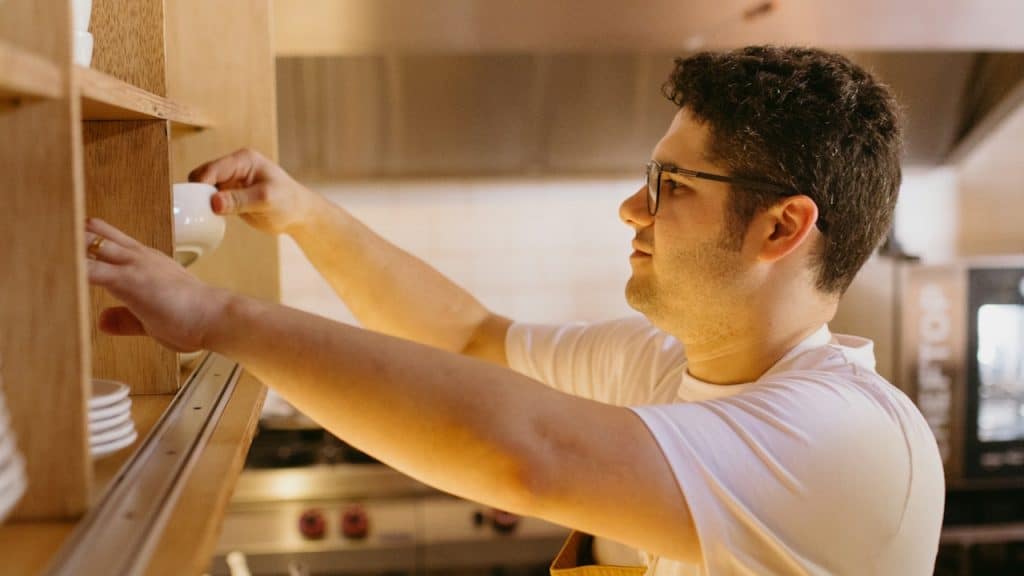 A man in glasses, a white shirt, and an apron is reaching up to place a small white cup on a wooden shelf in a commercial kitchen.