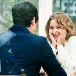 A woman smiling warmly, holding her face in her hands, looks across a table at a man.