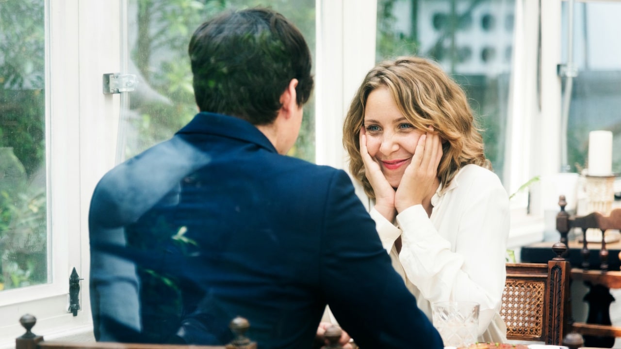 A woman smiling warmly, holding her face in her hands, looks across a table at a man.