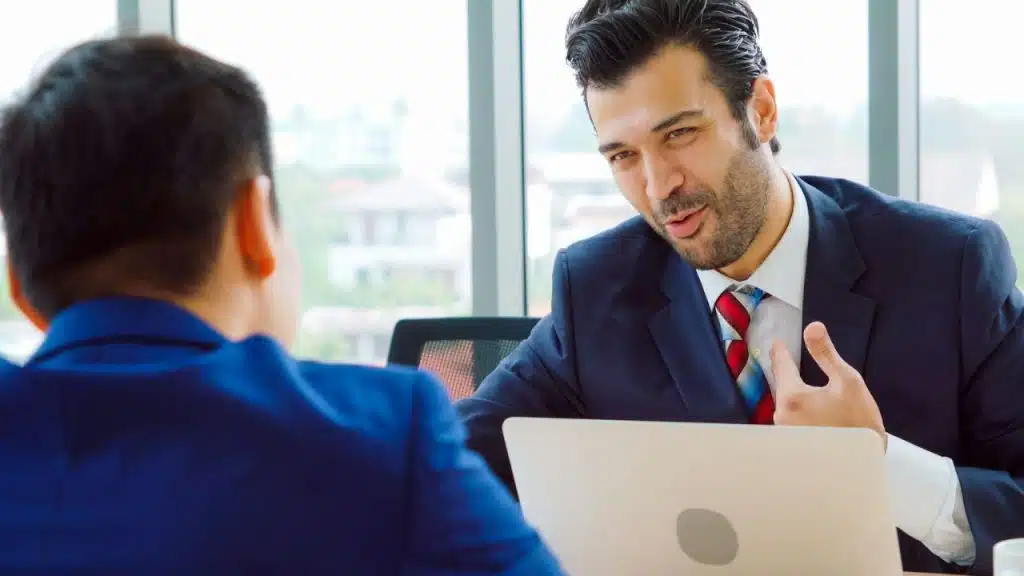 Two businessmen in suits talking across a laptop in a brightly lit office.