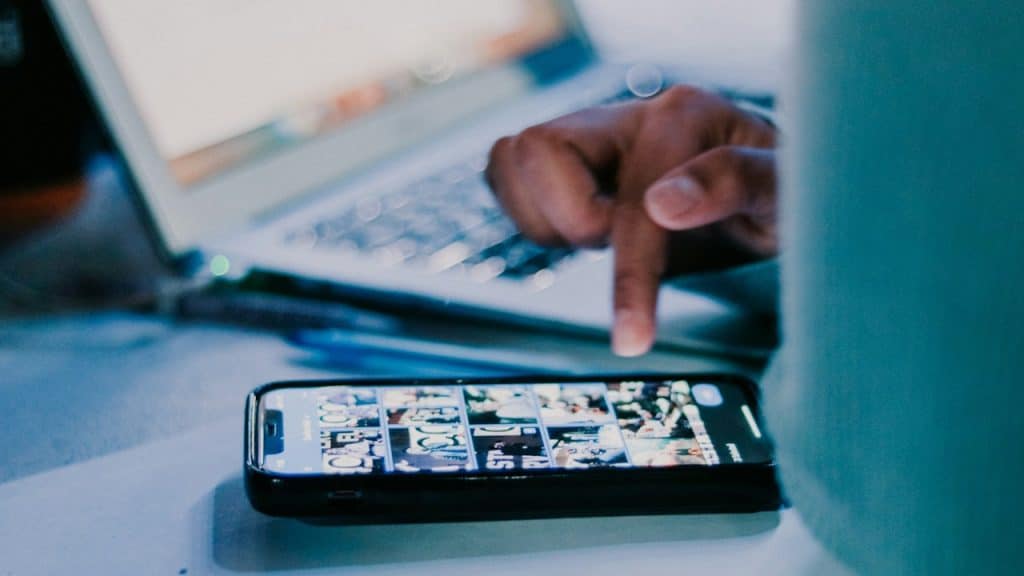 A close-up of a person's hand pointing at the screen of a smartphone lying on a desk, with an open laptop in the background.
