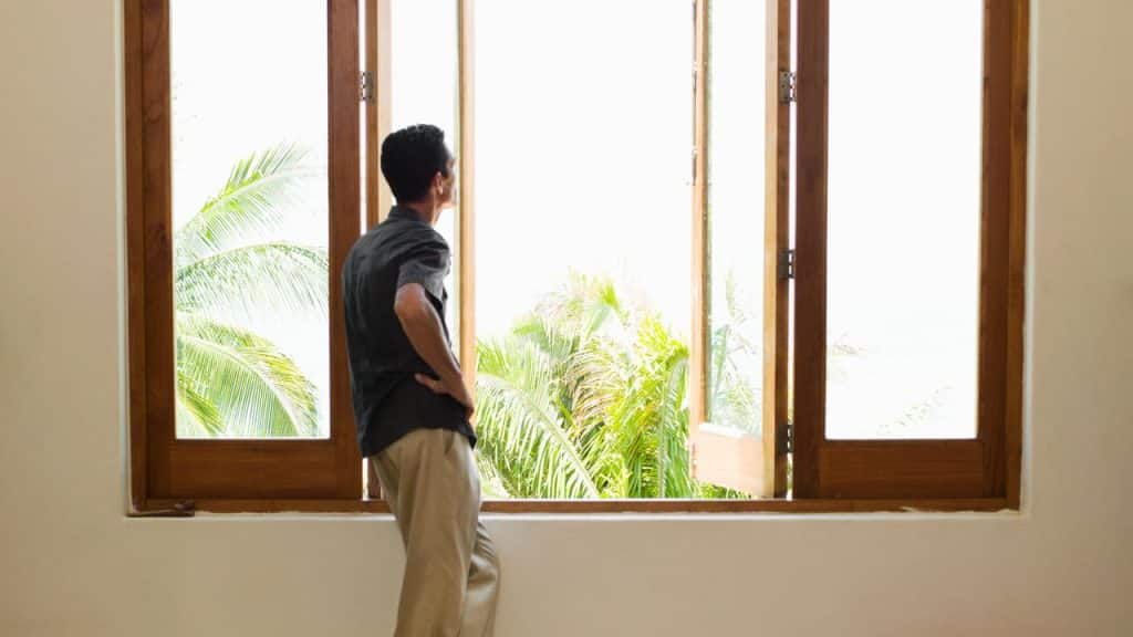 A man standing by an open window looking out at palm trees.