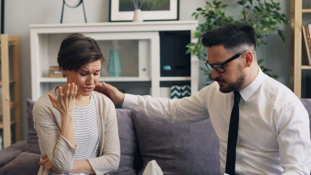 Man in a shirt and tie comforting a tearful woman on a sofa in a home setting.