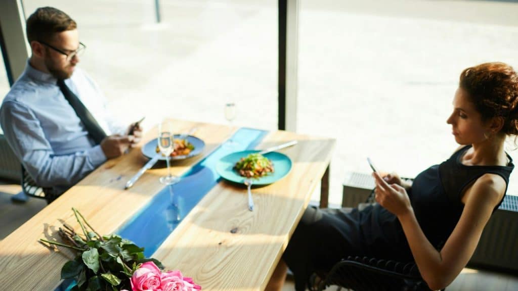 A man and woman sit opposite each other at a restaurant table, each looking at their phone.