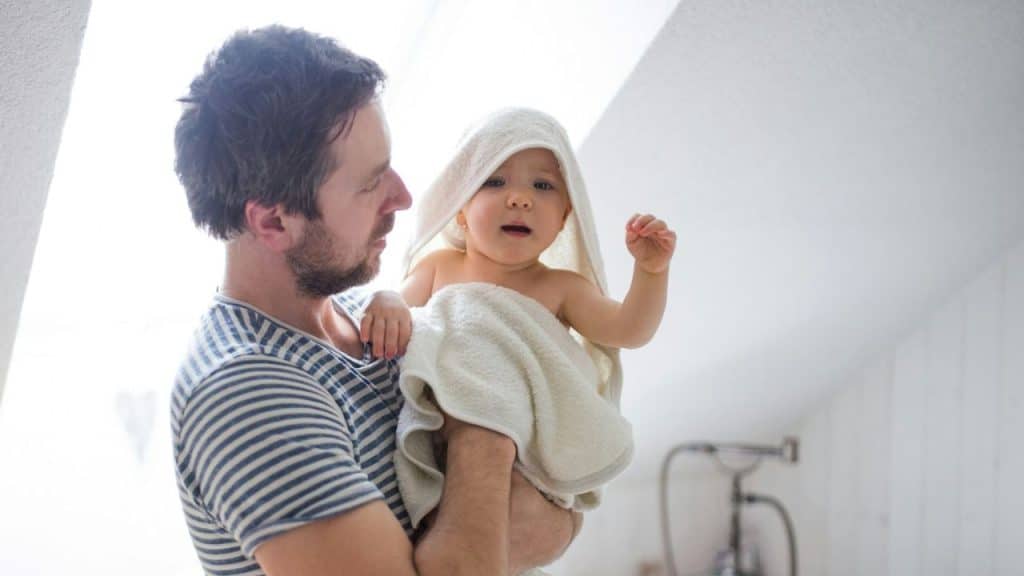 Man in striped shirt holding a baby wrapped in a white towel indoors.