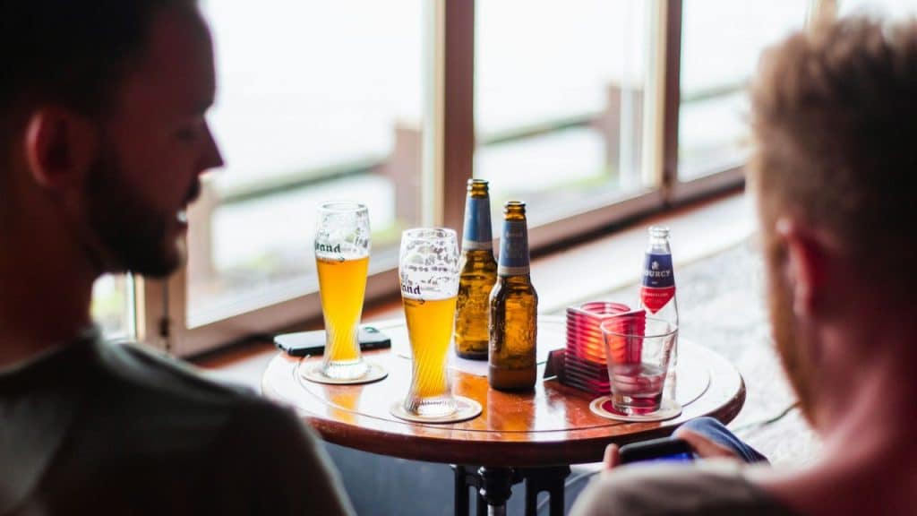 Two men sitting across a round table with beer glasses and bottles by a window.