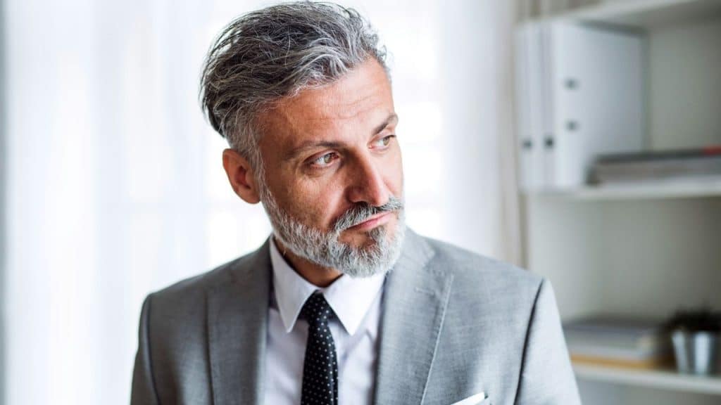 Man with gray hair and a beard wearing a suit and tie, looking away thoughtfully.