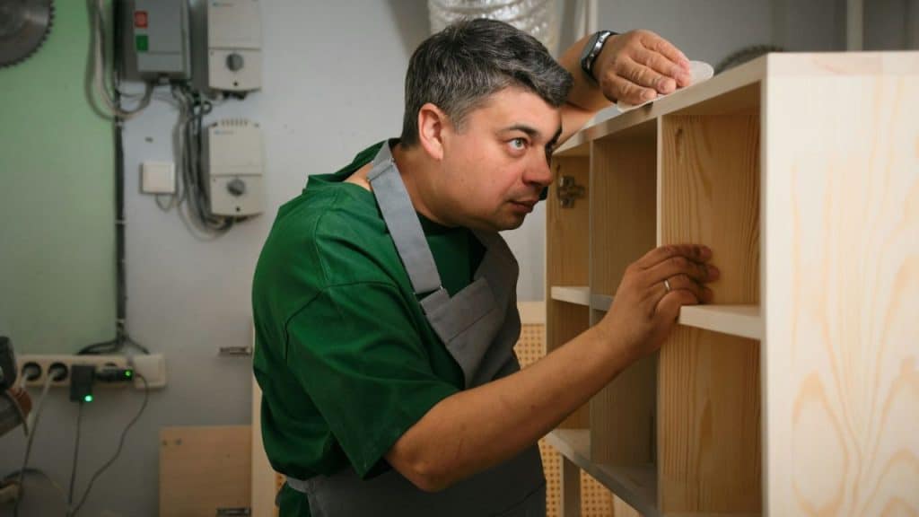 Man in a green shirt and apron examining wooden shelving in a workshop.