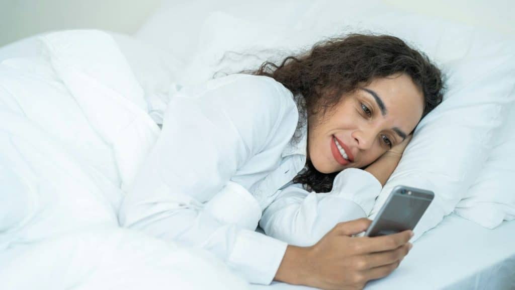Woman lying in a white bed, smiling while looking at a smartphone screen.