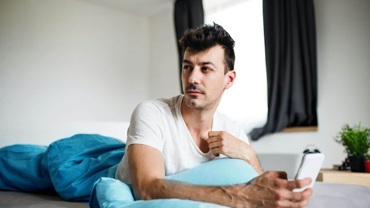 Man in a white shirt lying in bed with a blue blanket, holding a white smartphone.