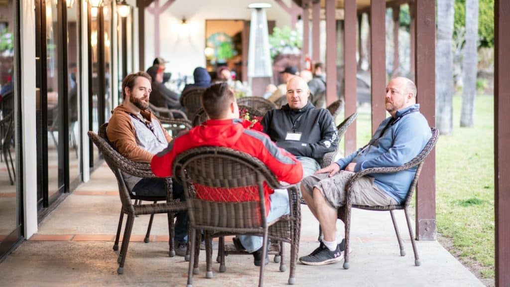 Four men sitting around an outdoor patio table talking at an event.