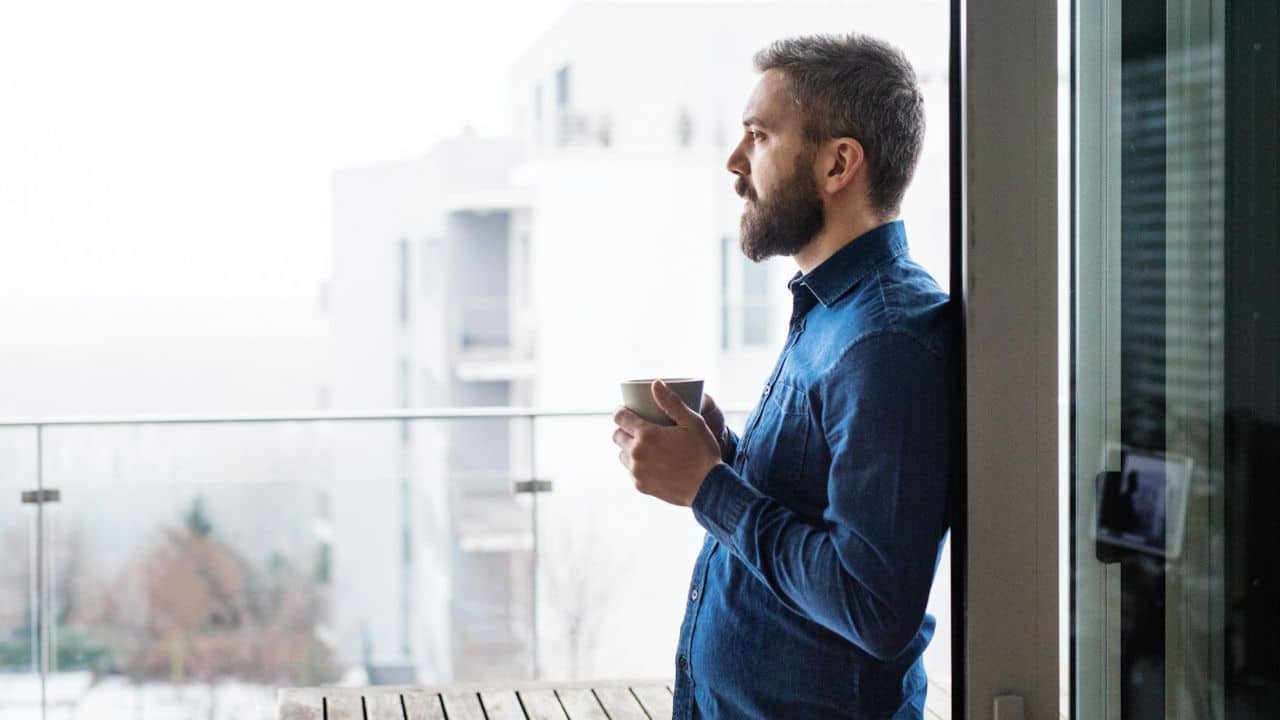 Bearded man in a blue shirt standing on a balcony, holding a mug and looking outside.