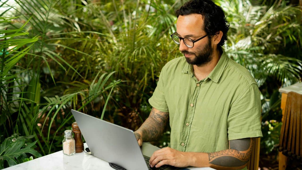 Man with glasses and tattooed arms working on a laptop in a lush garden setting.