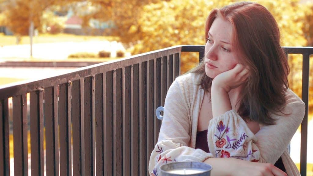 A young woman with reddish hair sits on a balcony, resting her chin on her hand, looking away.