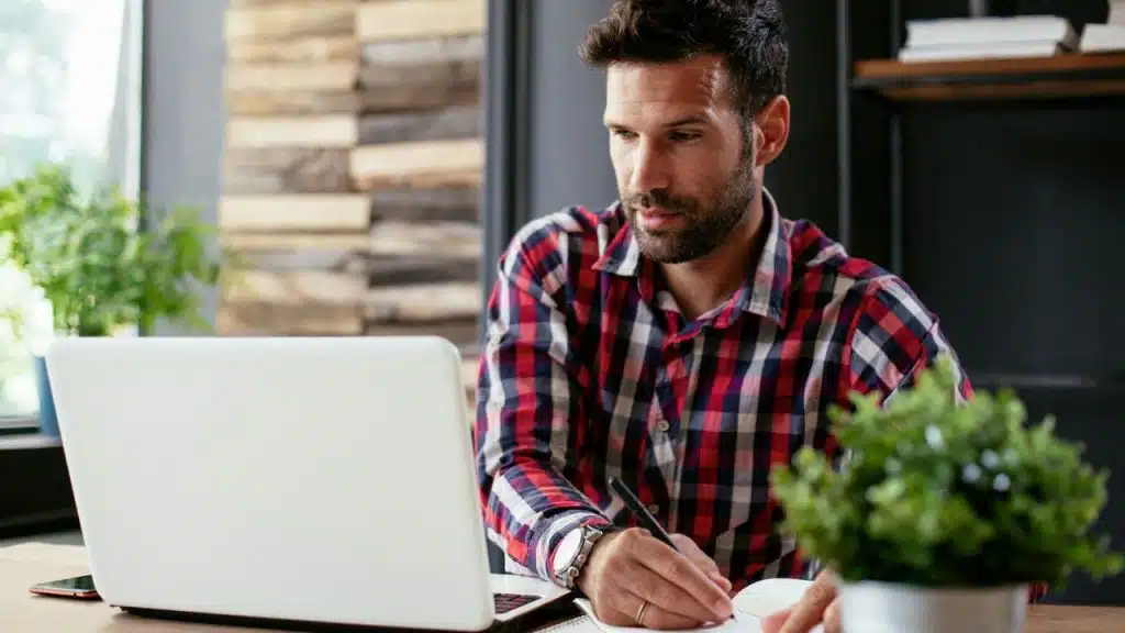 A man in a plaid shirt intently writes in a notebook while looking at a laptop.