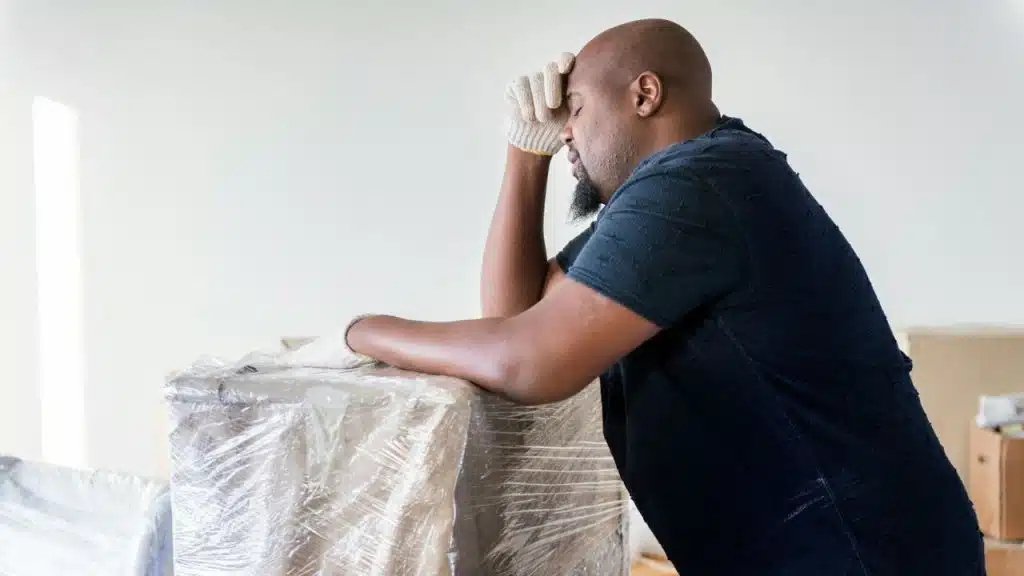A tired man wearing gloves leans on plastic-wrapped boxes in a bright room.