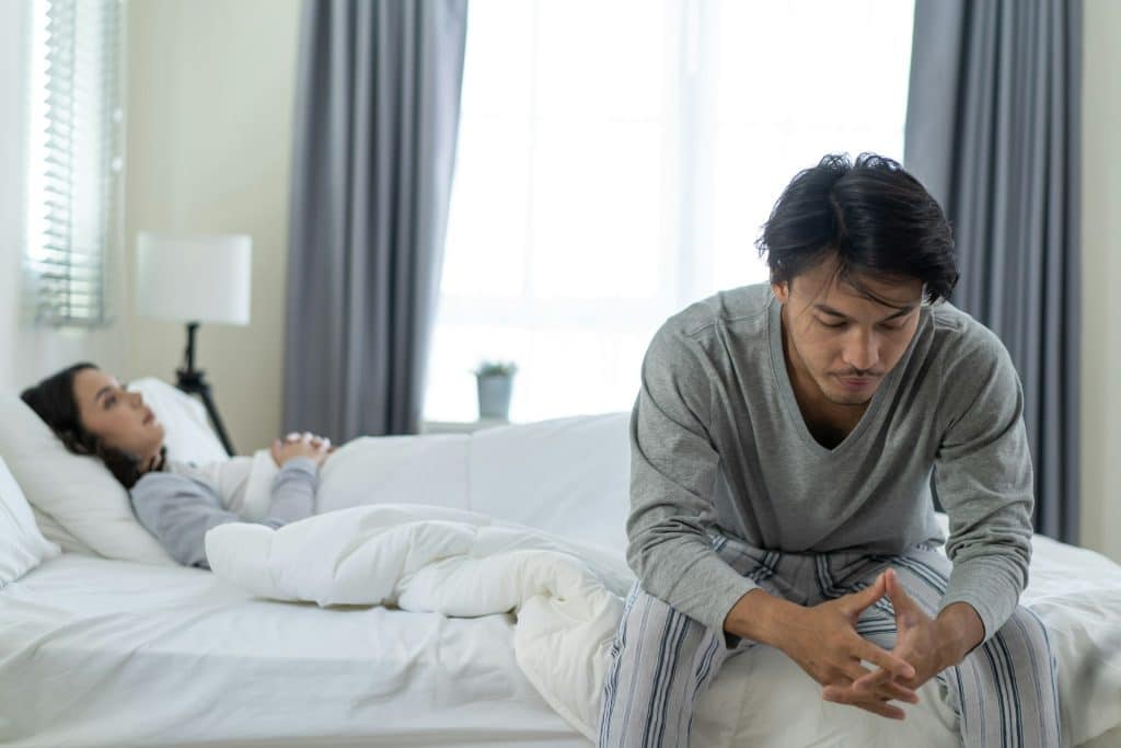 A man sitting on the edge of a bed looking worried while a woman lies behind him.