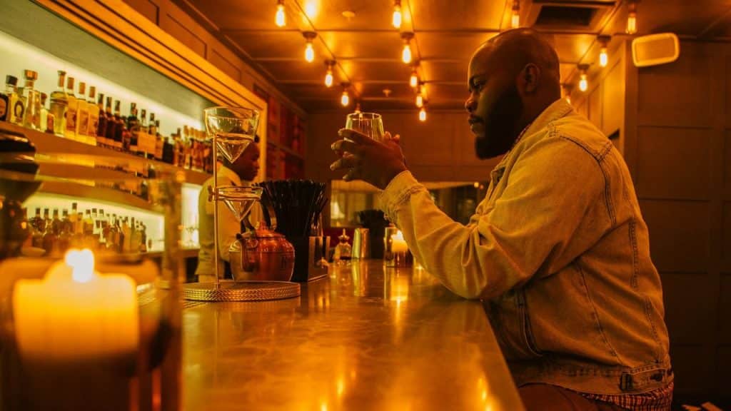 Man holding a glass at a warmly lit bar with many light bulbs.