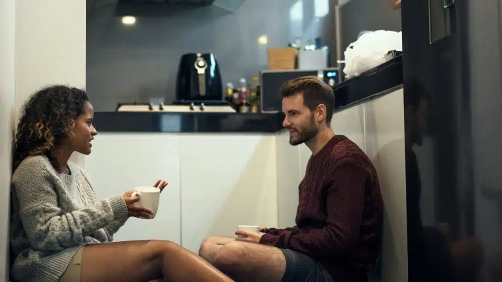 A man and woman are sitting on the kitchen floor talking and holding mugs.