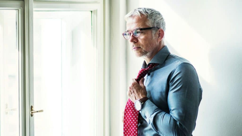Middle-aged man in a dress shirt and glasses loosening his red patterned tie by a window.