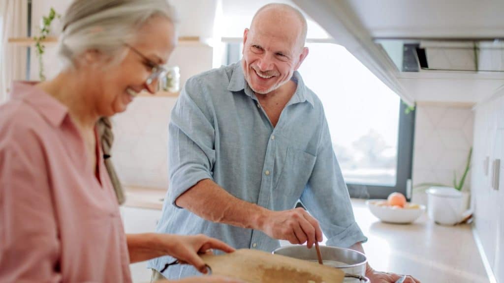 A smiling older man stirs a pot on the stove next to a woman holding a cutting board.