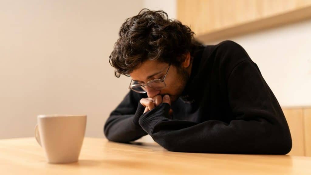 A person with glasses sits at a table, head down, hands clasped near a mug.