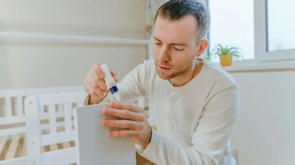 Man in a white shirt gluing a piece of white furniture in a bright room.